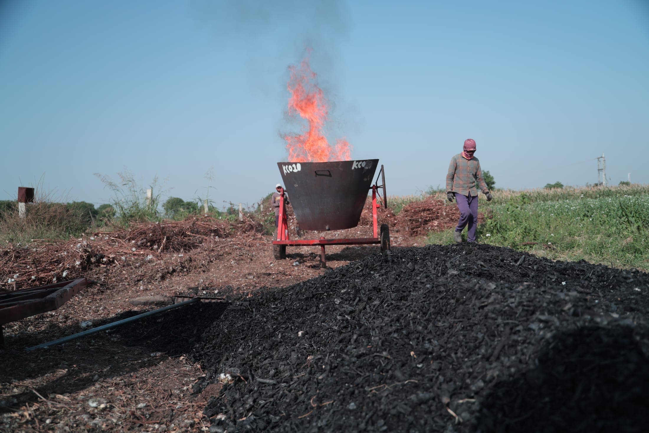 A pile of fresh biochar in the foreground with the kiln burning behind it