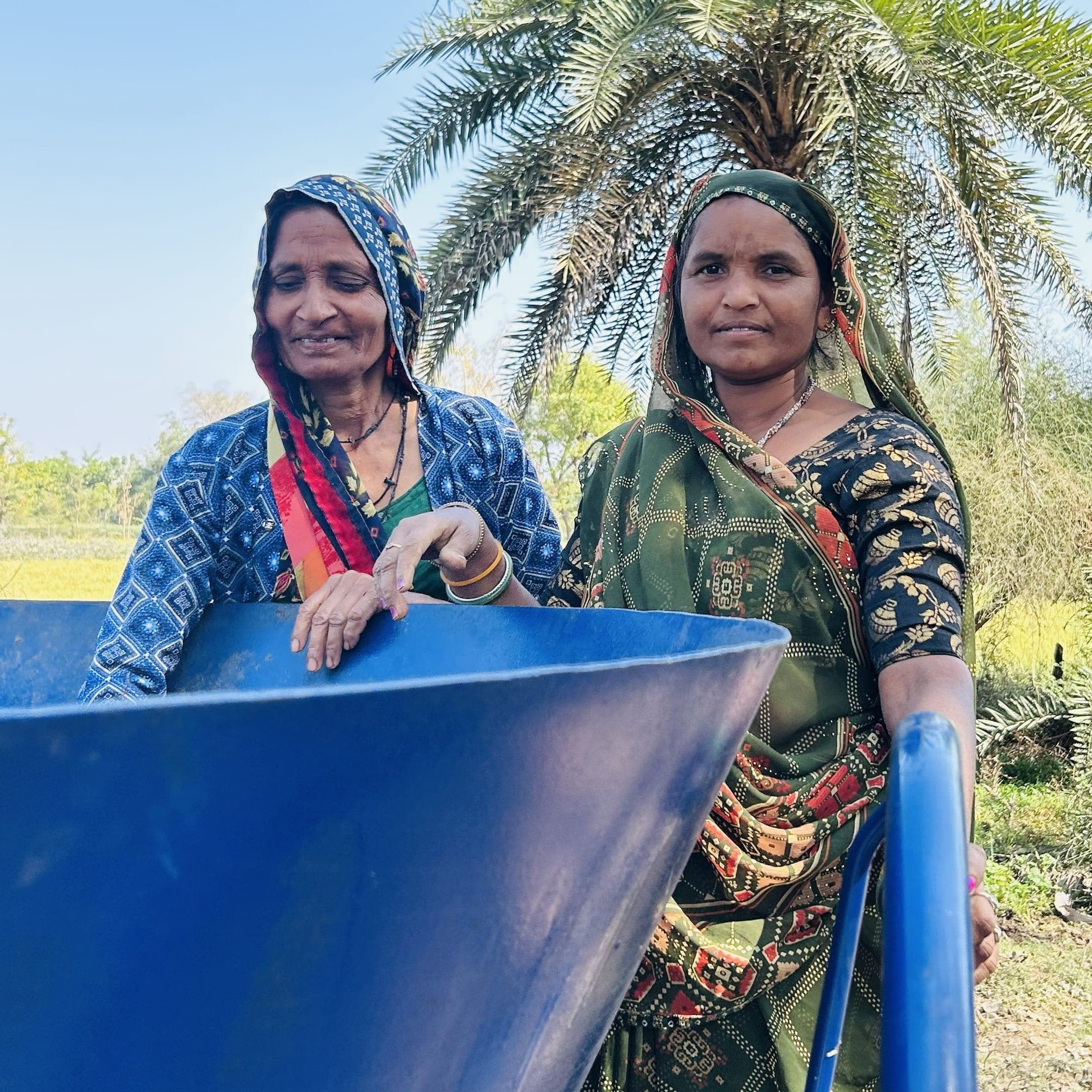Two women farmers stand smiling at a blue biochar kiln, a palm tree behind them