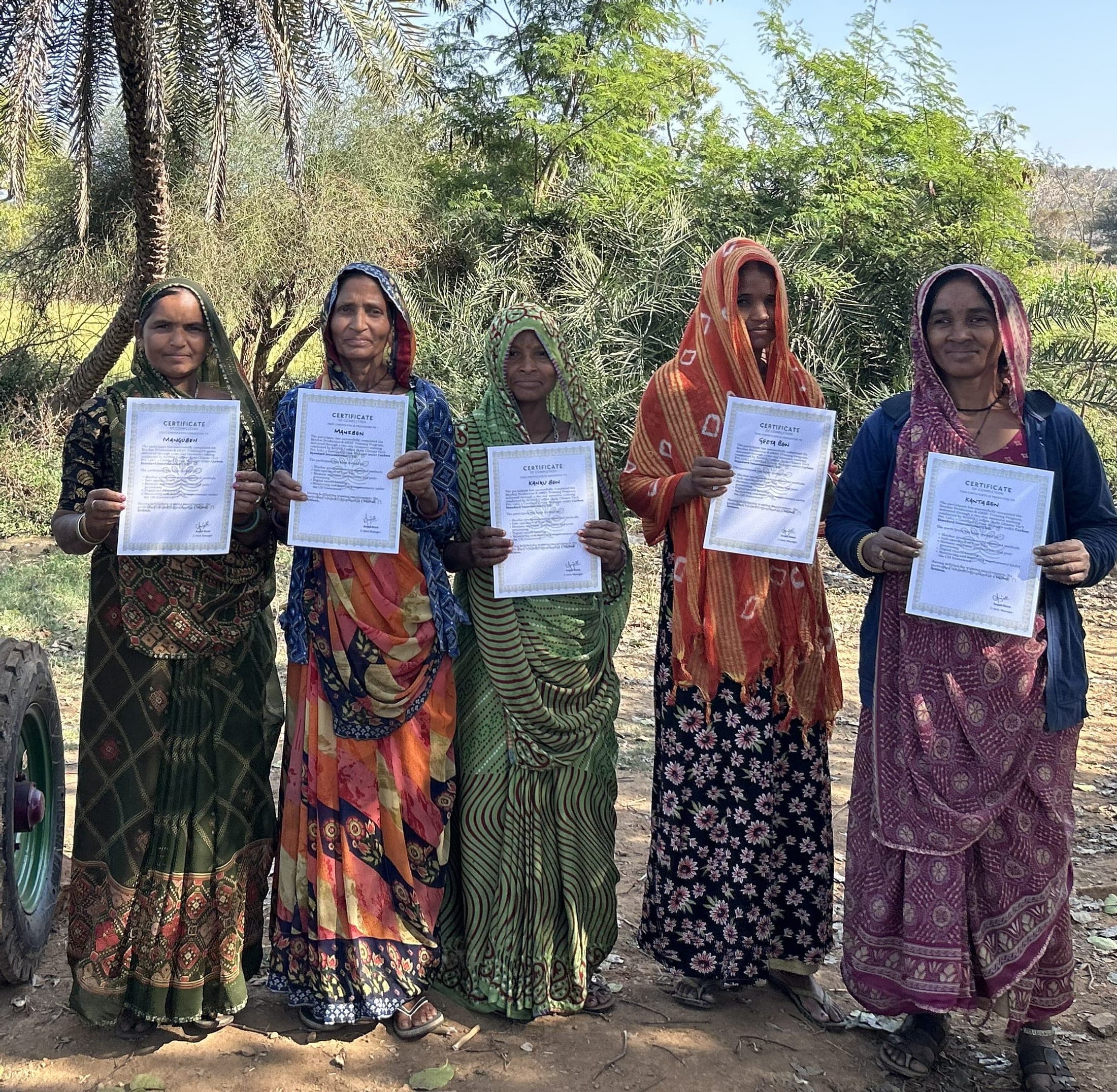 Five women farmers hold up their training certificates beside a biochar kiln