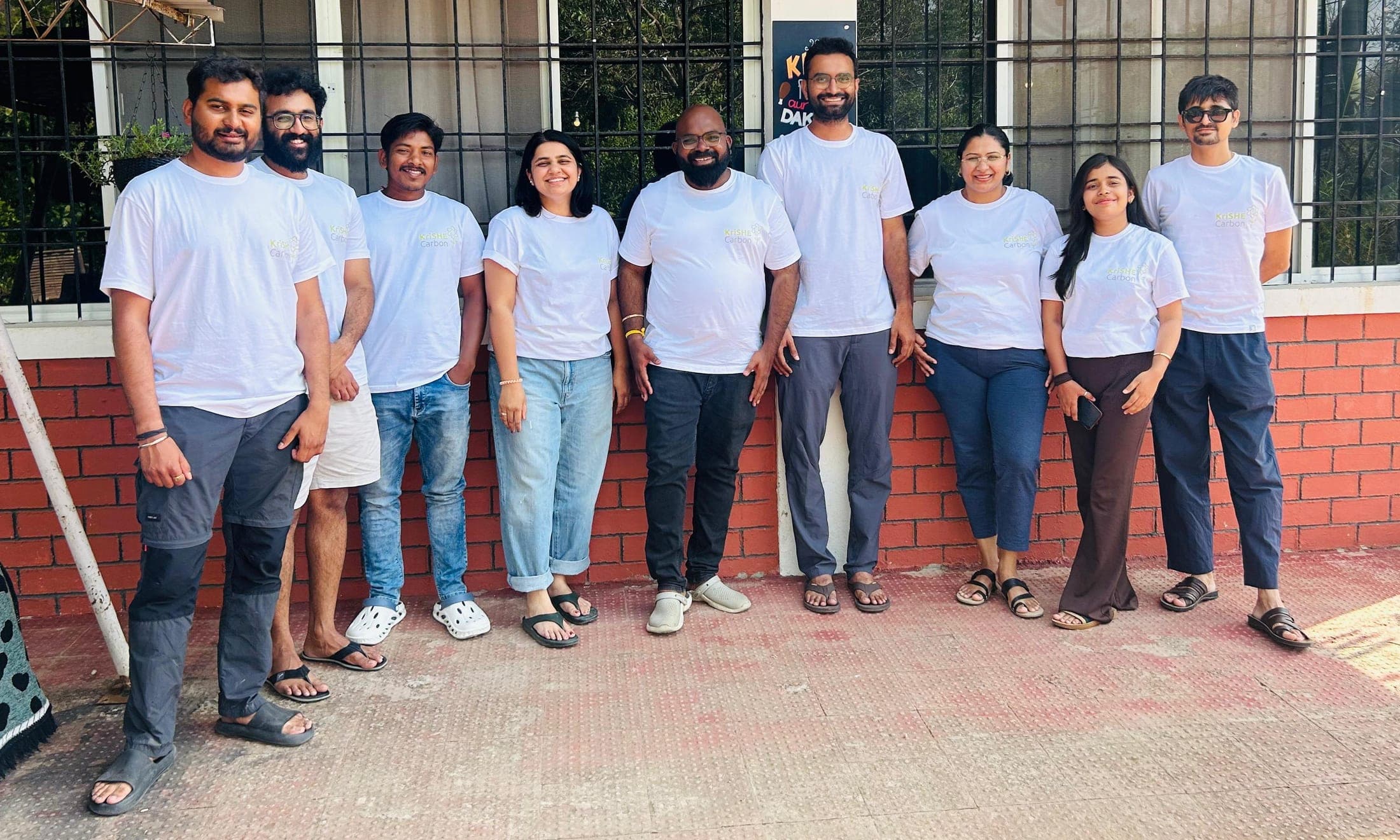 The KriSHE Carbon team: nine people in white T-shirts standing together outside a brick building
